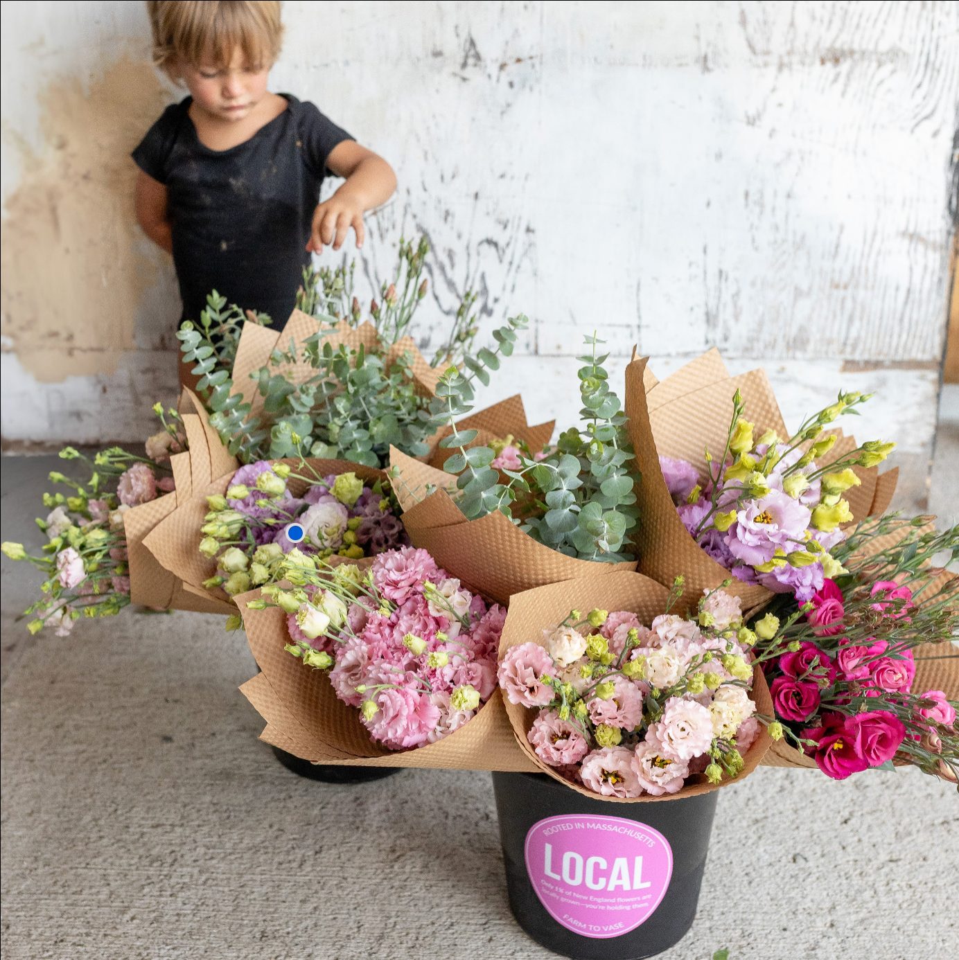 A child stands behind a black bucket filled with wrapped flower bouquets in a rustic indoor setting. The bouquets include pink, purple, and magenta flowers with green foliage, wrapped in brown paper, and the bucket has a pink label reading LOCAL.