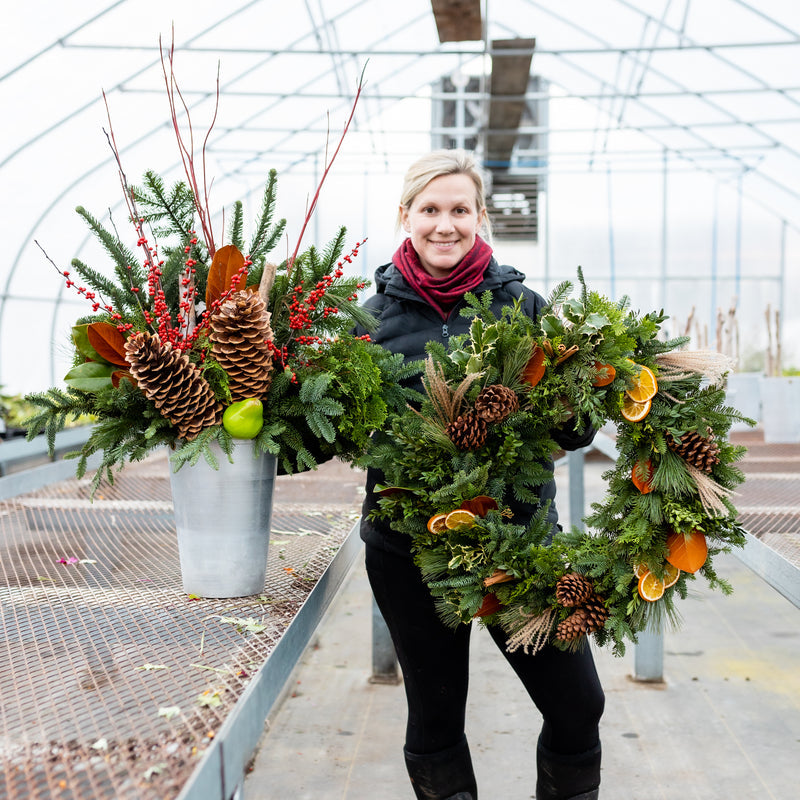 A floral designer holding an evergreen wreath beside a holiday planter with pinecones and red berries.