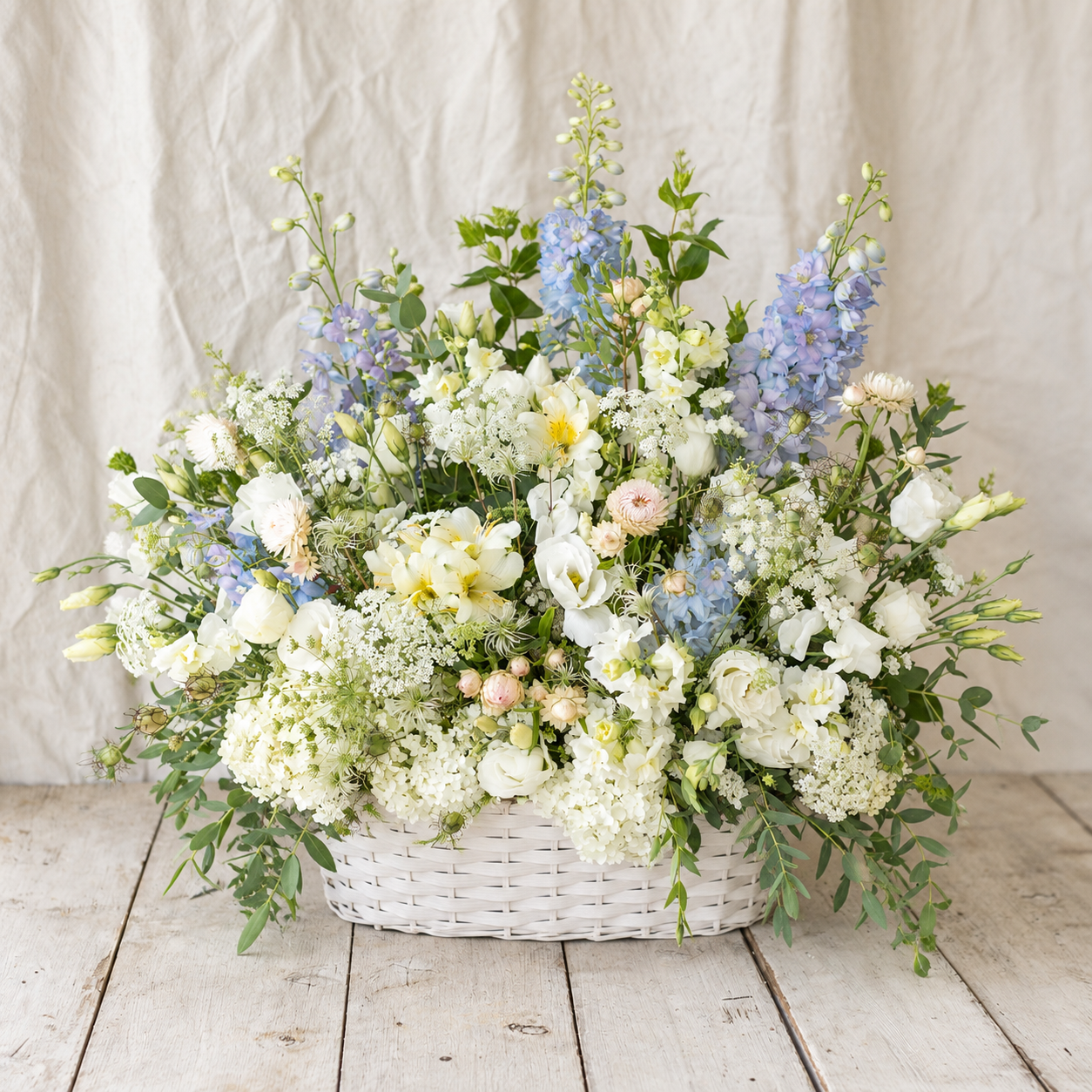 
                  
                    A large, lush floral arrangement in a white woven basket, featuring white and pale blue flowers with green foliage, set on a rustic wooden floor against a neutral fabric backdrop.
                  
                