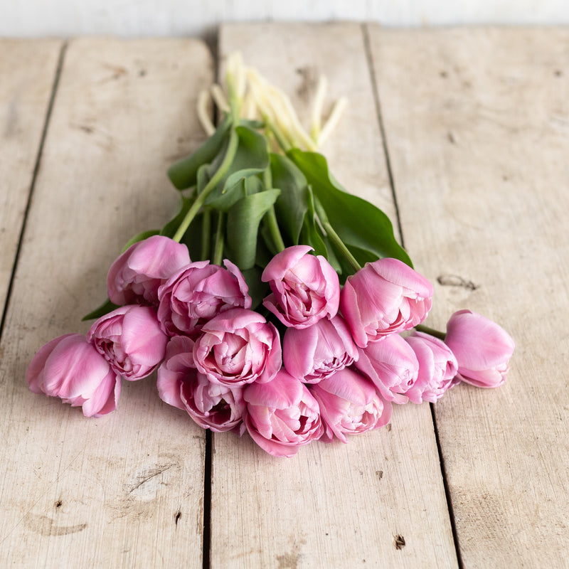
                  
                    Soft pink double tulips with layered petals resting on a rustic wooden table.
                  
                
